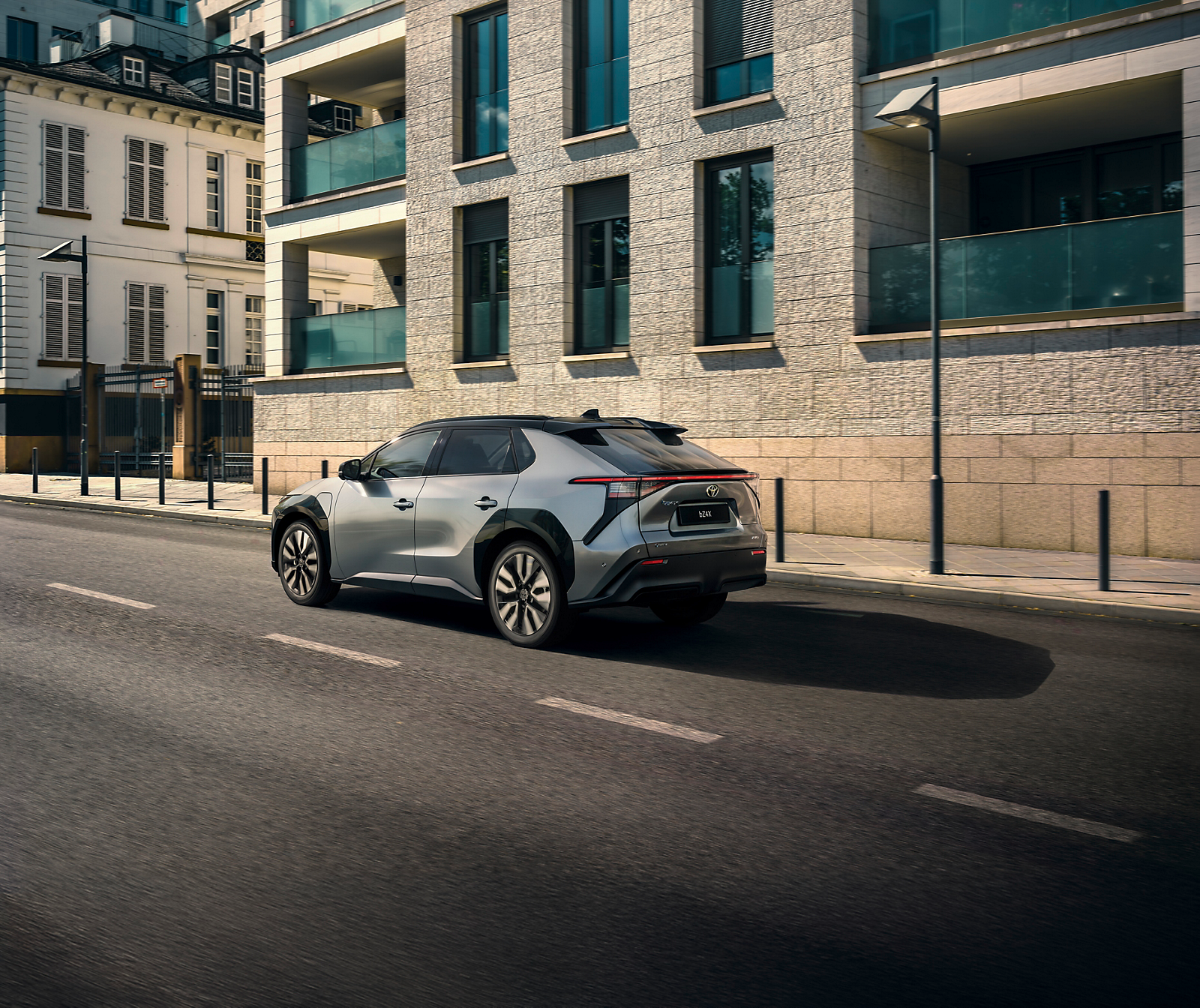 Ein silbergrauer Toyota bZ4X fährt auf einer Straße durch die Stadt, von hinten bei der Fahrt fotografiert, Gebäude im Hintergrund.