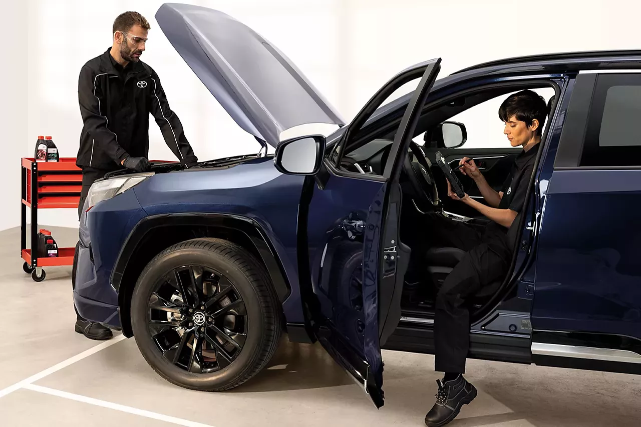 Zwei Toyota-Techniker bei der Wartung eines Urban Cruiser: Eine Person überprüft den Motorraum, die andere sitzt im Innenraum mit einem Diagnosetablet – professionelle Fahrzeugwartung im Autohaus, Service und Fehlerdiagnose am Hybridfahrzeug.